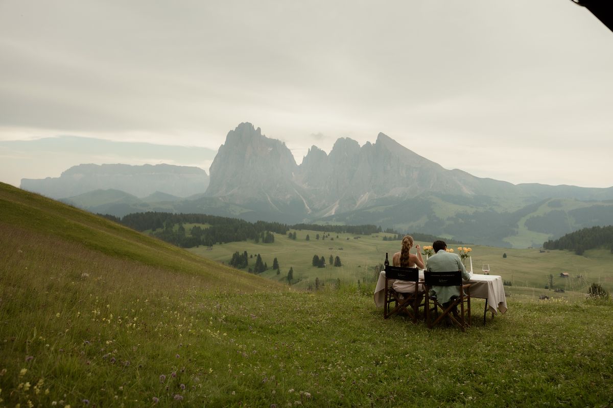 Paar beim Dinner an einem Tisch auf der Almwiese, dahinter Langkofel und Plattkofel